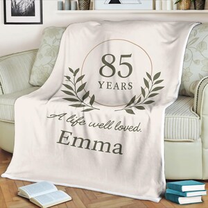 Könnte beinhalten: Cremefarbene Decke auf einem Sofa, mit dem Text "85 YEARS" in einem Kreis, umrahmt von Olivenzweigen. Die Decke trägt auch den Text "A life well loved. Emma."