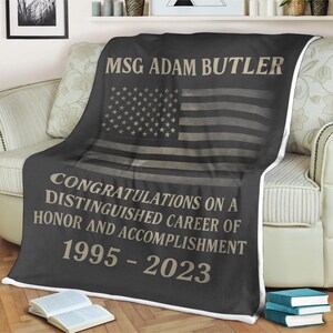 Puede incluir: Una manta de franela negra y gris con un borde blanco. La manta presenta un diseño de bandera estadounidense descolorida y el texto "MSG Adam Butler Felicitaciones por una distinguida carrera de honor y logro 1995-2023".
