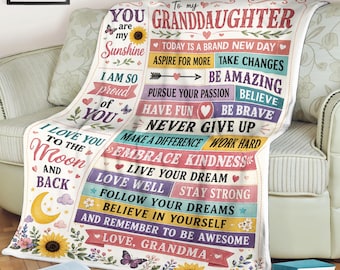May include: A white fleece blanket featuring the text To my GRANDDAUGHTER, YOU are my Sunshine, and I LOVE YOU TO THE MOON AND BACK. The design includes colorful horizontal bars with motivational phrases, yellow sunflowers, butterflies, and a crescent moon.