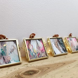May include: A row of five small, gold-framed pictures with abstract art. Each frame has a brown ribbon loop at the top. The artwork features a mix of colors, including pink, green, and gold, creating a modern aesthetic. The frames are displayed on a wooden surface.