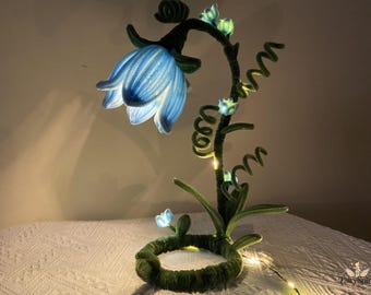 May include: A whimsical, handmade lamp shaped like a flower. The lamp features a large, light blue flower with illuminated petals, a green stem, and smaller glowing buds. The base is a green ring, and the lamp is lit with small fairy lights.