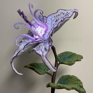 May include: A handcrafted, whimsical flower sculpture with a glowing centre. The large, textured petals are white with purple accents and black speckles. The stem is brown, and the leaves are green. The flower has a fantasy or fairy theme.