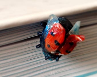 May include: A ladybug-shaped bookmark with red and black details, and a gold-colored, dome-shaped object. The bookmark is placed on the edge of a book. The ladybug has a blue body and transparent wings.