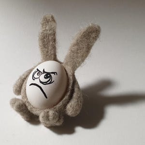 Könnte beinhalten: Eine skurrile Osterdekoration. Ein Ei mit einem wütenden Gesicht ist in einem grauen Filz-Hasenkörper mit langen Ohren und kleinen Pfoten eingebettet. Der Schatten des Hasen ist auf der weißen Oberfläche sichtbar.