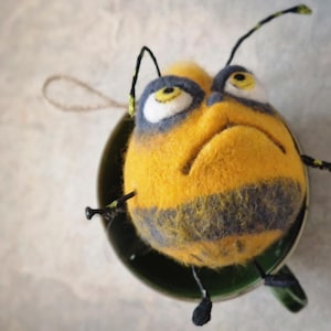 May include: A handcrafted felt bee decoration, featuring a yellow and black striped body, large eyes, and black antennae. The bee is placed in a green cup, creating a quirky decorative item. The ornament has a loop for hanging.