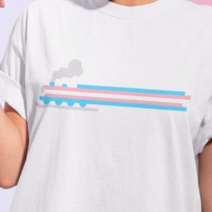 Puede incluir: Una camiseta blanca con un diseño de tren azul y rosa. El tren tiene una columna de humo gris y está en una vía con una franja azul y rosa.