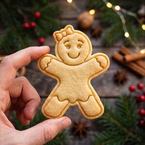 Pode incluir: Um biscoito de gengibre de cor castanha clara em forma de pessoa com um pequeno laço na cabeça. O biscoito apresenta detalhes faciais em relevo, incluindo olhos e uma boca sorridente, além de um design de borda recortada nos braços e pernas.