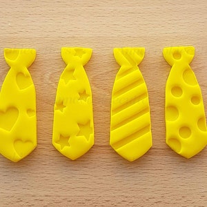 Op de afbeelding: Vier gele koekjessnijders in de vorm van een stropdas met reliëfdesigns: harten, sterren, strepen en cirkels. De uitstekers zijn van plastic en liggen op een houten oppervlak.