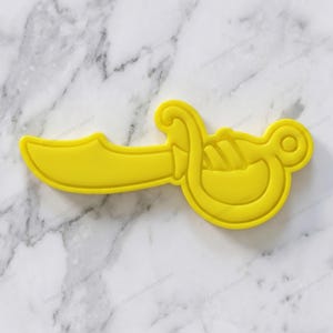 Op de afbeelding: Een gele plastic koekjessnijder in de vorm van een piratenzwaard. De snijder heeft een gebogen lemmet en een gedetailleerde handgreep met een cirkelvormige pommel. Het ontwerp is uit één stuk, geschikt voor het snijden van deeg.