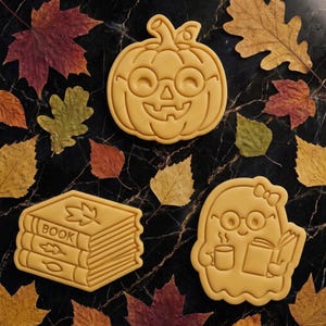 Può includere: Tre stampini per biscotti giallo chiaro raffiguranti una zucca con occhiali, una pila di tre libri con dettagli a foglia e la parola BOOK, e un fantasma con fiocco, occhiali, una tazza e un libro aperto, disposti tra varie foglie autunnali.