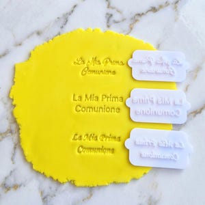 Può includere: Un impasto giallo brillante con il testo "La Mia Prima Comunione" impresso tre volte. Tre timbri rettangolari bianchi con lo stesso testo sono accanto all'impasto, con il testo specchiato sui timbri.