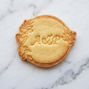 Peut inclure: Un biscuit rond de couleur beige clair présentant un motif en relief avec les lettres A et P séparées par une esperluette au centre. Le biscuit est orné d'un motif détaillé de feuilles et de vignes sur les bords gauche et droit du cercle.