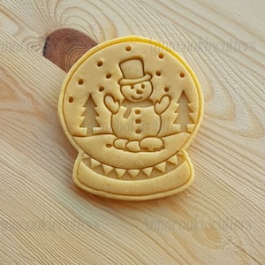 Könnte beinhalten: Ein hellbrauner Keks in Form einer Schneekugel mit einem Schneemann darin. Der Schneemann trägt einen schwarzen Hut und hat eine Karottennase. Auf beiden Seiten des Schneemanns befinden sich zwei kleine Bäume.