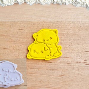 May include: Yellow plastic cookie cutter shaped like two cartoon cats hugging. The cat on top is holding the cat on the bottom.