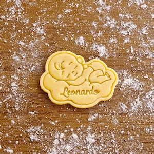 Può includere: Un tagliapasta giallo a forma di bambino che dorme con il nome "Leonardo" inciso su di esso. Il tagliapasta è su una superficie di legno ricoperta di polvere bianca.