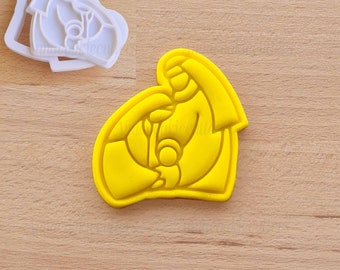 Puede incluir: Un cortador de galletas amarillo con forma de escena religiosa, con una madre y un niño. También hay un cortador de galletas blanco con el mismo diseño. Los cortadores son de plástico y están sobre una superficie de madera marrón claro.