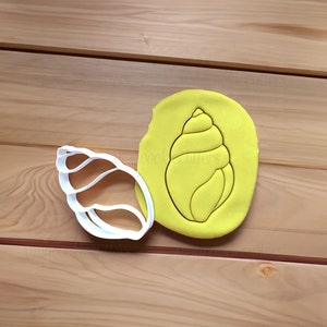 Könnte beinhalten: Ein weißer Plastik-Ausstecher in Form einer Muschel, neben einem gelben Stück Modelliermasse mit dem Abdruck der Muschel.