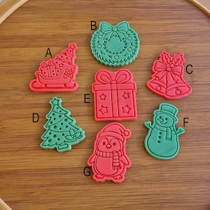Può includere: Una collezione di formine per biscotti a tema natalizio, rosse e verdi. Le forme includono una slitta, una ghirlanda, campane, un albero di Natale, un regalo, un pupazzo di neve e un pinguino. Disposti su una superficie di legno.