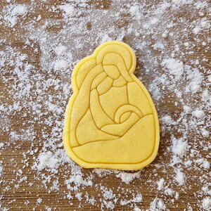 Può includere: Un tagliapasta giallo a forma della Sacra Famiglia, Maria, Giuseppe e il bambino Gesù. Il tagliapasta è su una superficie ricoperta di farina bianca.