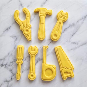 Può includere: Un set di sette tagliabiscotti in plastica gialla a forma di attrezzi da lavoro, tra cui pinze, martello, chiave inglese, cacciavite, chiave fissa, metro a nastro e sega a mano, disposti su due file.