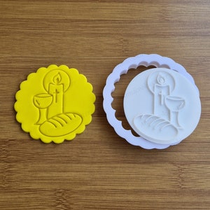 Può includere: Un tagliapasta in plastica gialla con un disegno di un calice, pane e una candela. Il tagliapasta è su una superficie di legno.