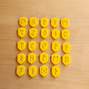 Op de afbeelding: Vierentwintig gele plastic runesteentjes met verschillende symbolen op elke tegel.