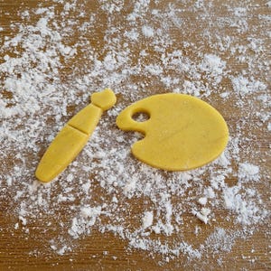 Può includere: Due stampini per biscotti gialli, uno a forma di pennello e l'altro a forma di tavolozza, sono su una superficie ricoperta di farina bianca.