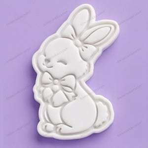 Puede incluir: Cortador de galletas de plástico blanco con forma de conejito con lazo en la cabeza y el pecho, sobre un fondo morado. El cortador tiene forma de conejito sonriente, ideal para hornear en Pascua o primavera.