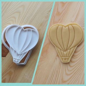 May include: A white plastic cookie cutter shaped like a heart-shaped hot air balloon. The cookie cutter is shown next to a cookie that has been cut out using the cookie cutter.