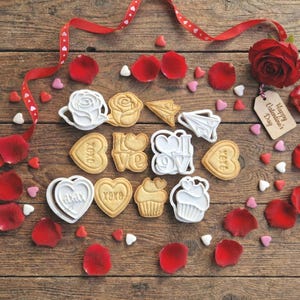 Pode incluir: Uma composição com tema de Dia dos Namorados com cortadores de biscoitos, biscoitos, pétalas de rosa e uma fita vermelha. Os cortadores de biscoitos têm a forma de rosas, corações e cupcakes. Os biscoitos dizem "Love" e "XOXO". Uma etiqueta diz "Happy Valentine's Day".