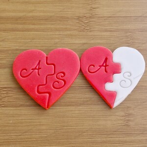 Può includere: Due biscotti a forma di cuore, uno rosso e uno bianco, con le lettere "A" e "S" su di essi. I biscotti sono progettati per incastrarsi come un puzzle.