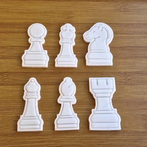 May include: Set of seven white plastic chess piece cookie cutters. The cutters include a pawn, a knight, a bishop, a rook, and a queen.