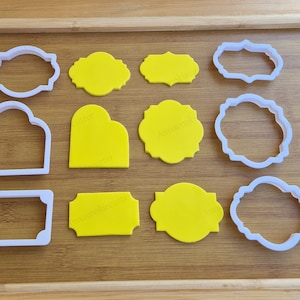 Op de afbeelding: Een set van 10 witte plastic koekjesvormen in verschillende vormen, waaronder rechthoeken, ovalen en harten. De vormen zijn gerangschikt op een houten oppervlak.