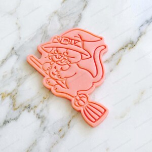 Può includere: Un tagliabiscotti rosa chiaro a forma di gatto con cappello da strega che cavalca una scopa. Il gatto tiene in mano una piccola zucca e il design presenta linee in rilievo per definire i tratti del viso, la fibbia del cappello e le setole.