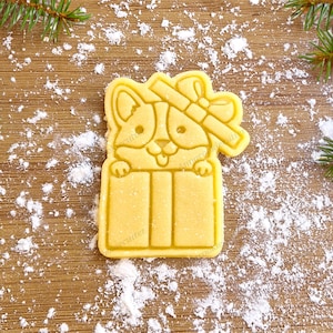 Può includere: Un tagliapasta giallo a forma di cane che spunta da una scatola regalo con un fiocco in cima. Il tagliapasta è su una superficie di legno ricoperta di polvere bianca.