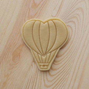 May include: A beige heart-shaped cookie cutter with a hot air balloon design. The balloon has a heart in the basket.