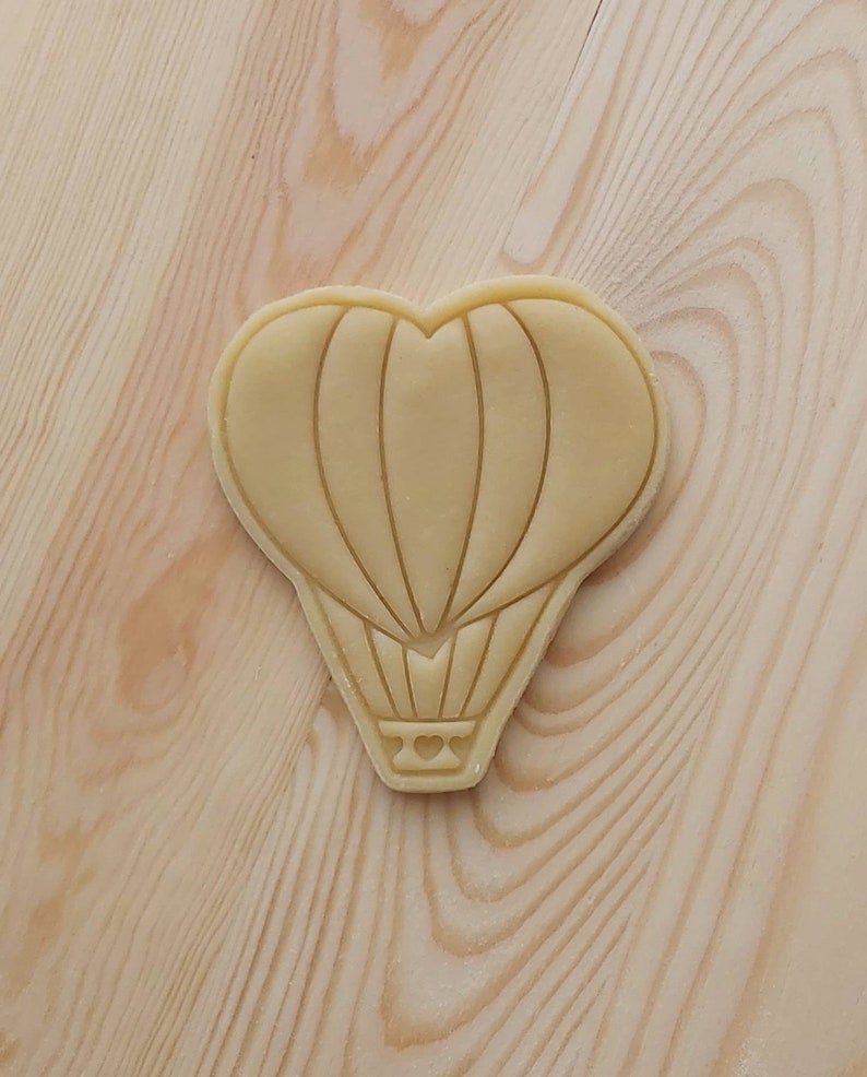 May include: A beige heart-shaped cookie cutter with a hot air balloon design. The balloon has a heart in the basket.