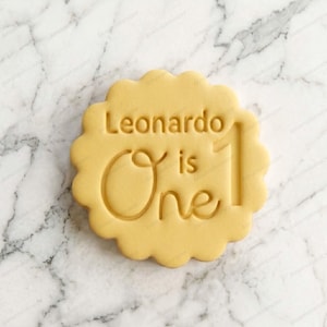 Può includere: Un tagliabiscotti rotondo con bordi smerlati realizzato in materiale giallo chiaro. Il centro presenta un testo in rilievo che recita Leonardo is One, con la parola One scritta in un grande carattere corsivo e il numero 1 posizionato a destra.