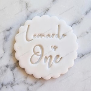 Può includere: Un tagliapasta bianco con bordo smerlato con le parole "Leonardo is One" in corsivo. Il tagliapasta è in plastica e poggia su una superficie di marmo. Ideale per fare biscotti per un primo compleanno.