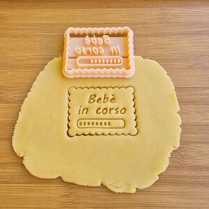 Può includere: Un tagliapasta di plastica rosa a forma di barra di avanzamento con il testo "Bebé in corso" e una barra di avanzamento. Il tagliapasta è sopra un pezzo rotondo di pasta per biscotti non cotta.