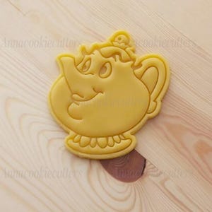 Może przedstawiać: Żółta foremka do ciastek w kształcie uśmiechniętego czajnika. Posiada uchwyt, dzióbek i dekoracyjną podstawę. Foremka leży na jasnej drewnianej powierzchni. Foremka do ciastek jest z tworzywa sztucznego.