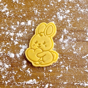 Può includere: Un biscotto di zucchero giallo a forma di coniglio che tiene un cuore. Il biscotto è su una superficie di legno marrone chiaro ricoperta di polvere bianca.
