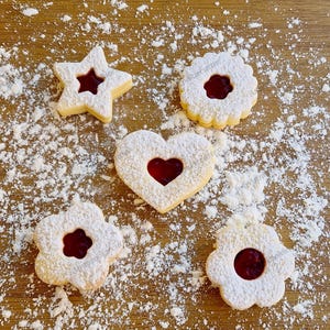Può includere: Vista dall'alto di cinque biscotti di pasta frolla a forma di stella, cuore, fiore e rotonda. Ogni biscotto ha un centro di marmellata rossa ed è spolverato con zucchero a velo. I biscotti sono disposti su una superficie di legno con zucchero a velo sparso.