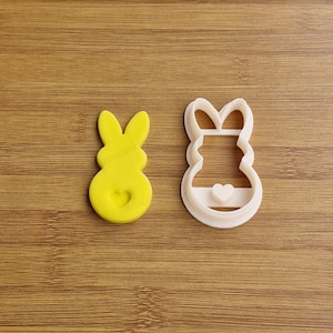 Può includere: Un tagliapasta di plastica bianca a forma di coniglio con un cuore sul ventre. Il tagliapasta è accanto a un biscotto di coniglio di pasta di zucchero giallo.