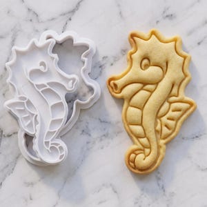 May include: A white plastic seahorse cookie cutter positioned next to a baked, light tan seahorse-shaped cookie. The cutter features a detailed outline and interior impressions that match the embossed design on the baked cookie surface.