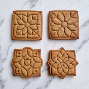 Könnte beinhalten: Vier hellbraune, quadratische und geformte Kekse mit geprägten Blumenmustern. Zwei Kekse sind einfache Quadrate mit einem zentralen Blumenmotiv, während die anderen beiden dekorative Ränder mit geometrischen Blumenmustern und kleinen Vertiefungen aufweisen.