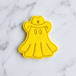 Può includere: Un tagliabiscotti giallo brillante a forma di fantasma che indossa un cappello da cowboy. Il design presenta una stella centrale sul cappello, due ritagli ovali per gli occhi e un bordo inferiore fluido e drappeggiato che imita un fantasma.