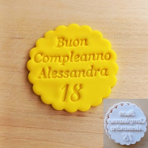 Puede incluir: Galletas de fondant amarillo con el texto "Buon Compleanno Alessandra 18" en letras blancas. Un cortador de galletas de plástico blanco con el mismo texto se muestra en el fondo.