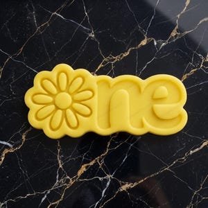 May include: A yellow plastic cookie cutter shaped like a daisy flower followed by the lowercase letters o, n, and e, forming the word one. The cutter has a raised border and a flat base, designed for cutting dough into a specific shape.