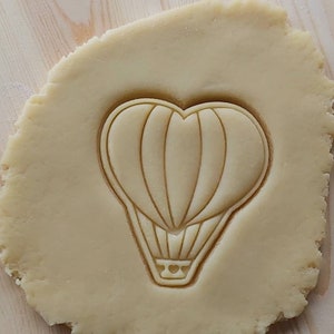 May include: A close-up of a cookie dough sheet with a hot air balloon cookie cutter pressed into it. The cookie cutter is shaped like a heart with a basket at the bottom.